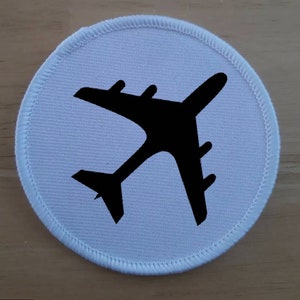 May include: White round fabric patch with a black airplane silhouette in the centre.