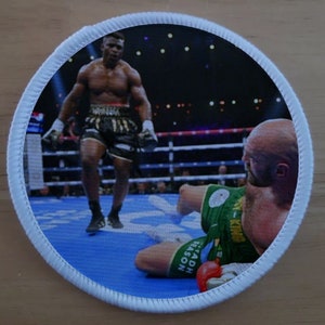 May include: A round, white-bordered patch with a boxing scene. A black boxer in black shorts and white gloves stands over a white boxer in green shorts who is lying on the blue mat. The white boxer is wearing a red boxing glove. The text "SADH EASON" is visible on the white boxer's shorts.