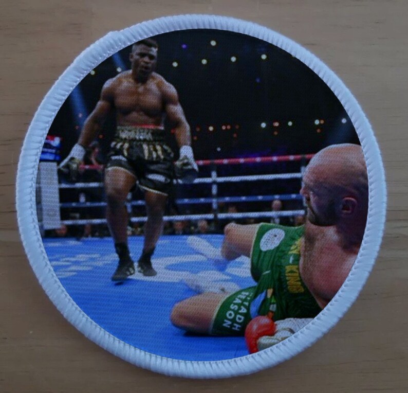 K&ouml;nnte beinhalten: Ein runder, wei&szlig; umrandeter Patch mit einer Boxszene. Ein schwarzer Boxer in schwarzen Shorts und wei&szlig;en Handschuhen steht &uuml;ber einem wei&szlig;en Boxer in gr&uuml;nen Shorts, der auf der blauen Matte liegt. Der wei&szlig;e Boxer tr&auml;gt einen roten Boxhandschuh. Der Text "SADH EASON" ist auf der Shorts des wei&szlig;en Boxers sichtbar.