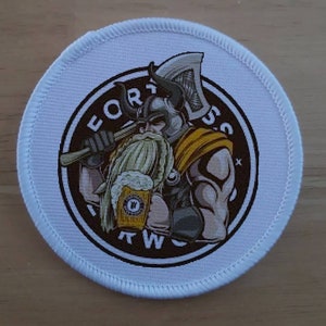 May include: White round patch with a brown circle in the centre. The circle features a cartoon viking holding a mug of beer and an axe. The text "FORTRESS TWO" is written around the circle.