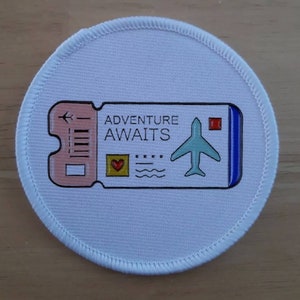 May include: A white round patch with a pink and blue airplane ticket design. The ticket says "Adventure Awaits" and has a small red heart and a blue airplane.