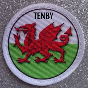 Peut inclure: Un écusson rond avec le mot "TENBY" en haut. Il présente un dragon rouge, symbole du Pays de Galles, sur un fond blanc et vert, avec une bordure blanche. L'écusson est un souvenir.