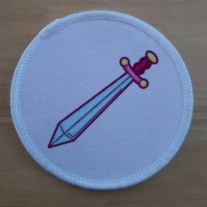Può includere: Toppa rotonda in tessuto bianco con un design a spada rosa e blu.