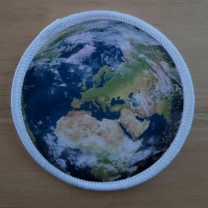 May include: A white fabric patch with a blue and green image of the Earth. The image shows the continents and oceans of the world.