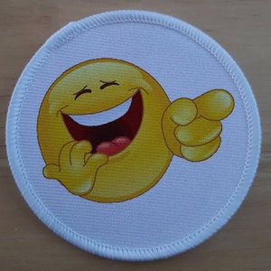 May include: A yellow emoticon with a big smile and a closed eye, laughing and pointing to the right. The emoticon is on a white circular patch.