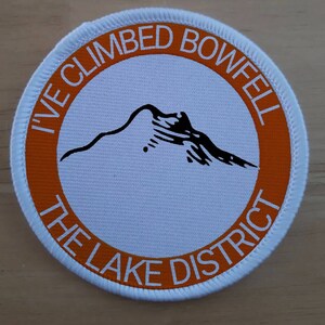 May include: White round patch with an orange border. The text "I've Climbed Bowfell The Lake District" is printed in a white circle on the patch. A black silhouette of a mountain is in the center of the circle.
