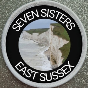 May include: A white and black round patch with the text "SEVEN SISTERS EAST SUSSEX" in a white circle on a black background. The centre of the patch features a photo of the Seven Sisters Cliffs in East Sussex, England.