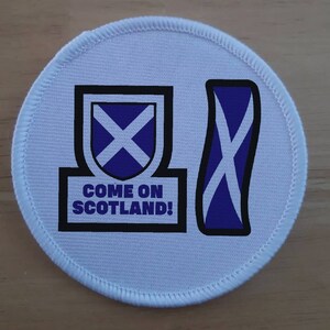 Peut inclure: Écusson rond blanc avec un motif de drapeau écossais bleu et blanc. Le texte "COME ON SCOTLAND!" est imprimé sur l'écusson.