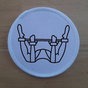 May include: White round patch with a black line drawing of a person with their arms and legs bound, hanging upside down from a bar.