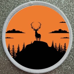 Puede incluir: Una silueta negra de un ciervo de pie en la cima de una montaña contra un atardecer naranja con nubes negras. El ciervo tiene cuernos y mira hacia adelante. Hay árboles en primer plano.