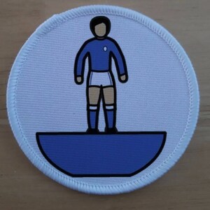 Könnte beinhalten: Ein weißer Stoff-Patch mit einem blauen und weißen Cartoon-Bild eines Fußballspielers, der auf einem blauen Halbkreis steht.