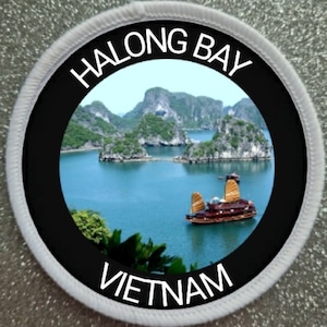 Può includere: Una toppa circolare bianca con bordo nero e un cerchio nero al centro. Il cerchio nero contiene una foto di una baia con isole verdi e una barca tradizionale vietnamita. Il testo "HALONG BAY VIETNAM" è stampato in bianco attorno al cerchio nero.