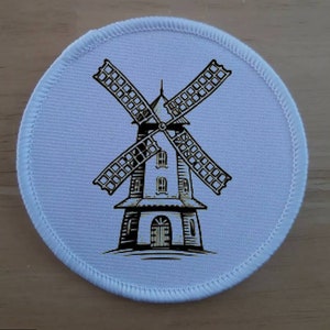 Op de afbeelding: Een ronde, witte patch met een gedetailleerde illustratie van een windmolen in bruin- en zwarttinten. De windmolen heeft vier wieken en een meerlaagse structuur. De patch heeft een gestikte rand.
