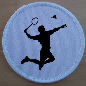 Può includere: Una toppa rotonda bianca con una silhouette nera di una persona che gioca a badminton. La persona sta saltando e oscillando una racchetta per colpire un volano. La toppa ha un bordo cucito.