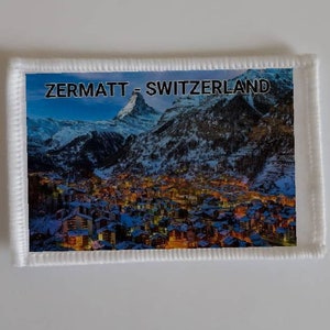 Könnte beinhalten: Ein weißer Patch mit einer genähten Umrandung, der ein Foto eines schneebedeckten Bergdorfes mit dem Text "ZERMATT - SCHWEIZ" oben zeigt.