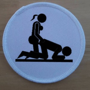 May include: A white, circular patch with a black graphic depicting two stick figures in a sexual position. The patch has a white border with a stitched edge. The graphic is centered on the patch.