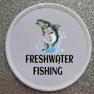 Może przedstawiać: Okrągła biała łatka z czarną obwódką i grafiką skaczącej ryby. Tekst "FRESHWATER FISHING" jest nadrukowany czarnym kolorem pod rybą.