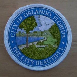 May include: A white, round patch with blue trim and a white background. The patch features a green palm tree, a blue lake, and a green path. The text "City of Orlando, Florida" and "The City Beautiful" are printed in white around the edge of the patch.
