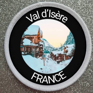 Könnte beinhalten: Ein runder weißer Patch mit schwarzem Text und einem schwarzen Kreis, der ein Foto eines verschneiten Dorfes in den französischen Alpen enthält. Der Text auf dem Patch lautet "Val d'Isère FRANCE".