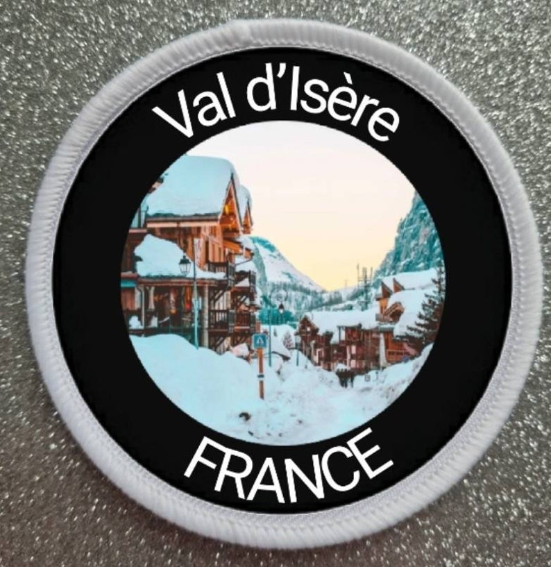 Op de afbeelding: Een ronde witte patch met zwarte tekst en een zwarte cirkel met een foto van een besneeuwd dorp in de Franse Alpen. De tekst op de patch luidt "Val d'Is&egrave;re FRANCE".