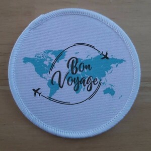 May include: A white round fabric patch with a blue world map design and the text "Bon Voyage" in black cursive font. Two small black aeroplanes are flying above the map.