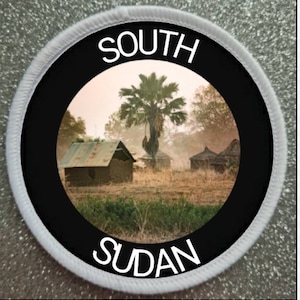 May include: A round, white patch with a black circle in the center. The circle contains a photo of a village in South Sudan with a palm tree in the foreground. The text "SOUTH SUDAN" is printed in white on the patch.