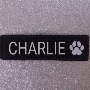 May include: Black fabric dog tag with white text that reads "CHARLIE" and a white paw print.
