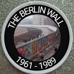 Könnte beinhalten: Ein schwarzer und weißer runder Patch mit dem Text "THE BERLIN WALL 1961 - 1989" am Rand. Die Mitte des Patches zeigt ein Farbfoto der Berliner Mauer mit Graffiti.