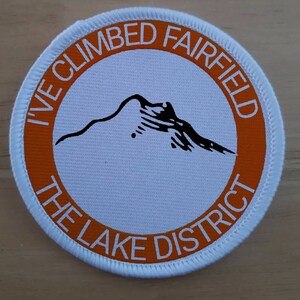 May include: White round patch with an orange border. The text "I've Climbed Fairfield The Lake District" is printed in a white circle on the orange border. A black silhouette of a mountain is in the center of the patch.