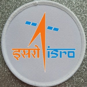 Könnte beinhalten: Weißer runder Patch mit einer orangefarbenen Rakete und zwei blauen Satelliten. Der Text "इसरो isro" ist in Blau und Schwarz gedruckt.