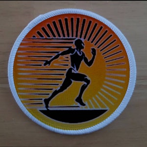 Puede incluir: Un parche redondo con borde blanco que muestra la silueta negra de un corredor sobre un fondo de sol amarillo y naranja.