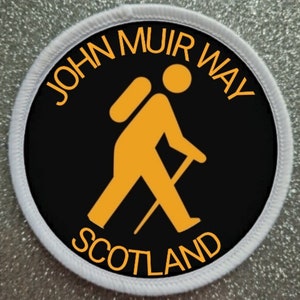 May include: A black and white round patch with white stitching around the edge. The patch features a yellow silhouette of a hiker with a backpack and walking stick. The text "JOHN MUIR WAY SCOTLAND" is written in yellow around the hiker.