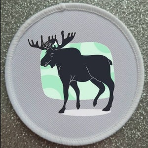 May include: A white round patch with a black silhouette of a moose with large antlers standing on a green and white background.