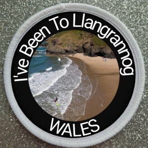 Op de afbeelding: Een ronde witte patch met zwarte tekst die "I've Been To Llangrannog WALES" luidt. De patch heeft een foto van een strand met golven en een zanderige kust.