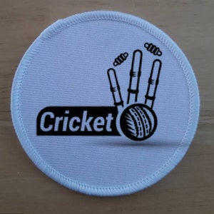 May include: White round fabric patch with black stitching around the edge. The patch features a black and white illustration of a cricket bat and ball with the word "Cricket" printed in black.