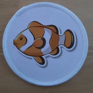 May include: A white round fabric patch with a cartoon image of an orange and white clownfish. The fish has black eyes and a black outline.