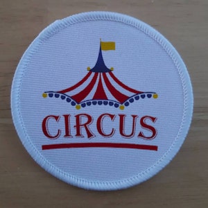 Könnte beinhalten: Runder Stoff-Patch in Weiß mit einem rot-weiß-blauen Zirkuszelt mit einer gelben Flagge oben. Das Wort "CIRCUS" ist in Rot unter dem Zelt aufgedruckt.