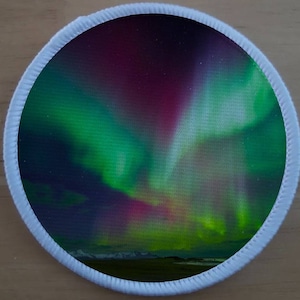 Puede incluir: Un parche redondo con un borde blanco y una imagen colorida de la aurora boreal, un espectáculo de luz natural en el cielo, predominantemente verde y morado.