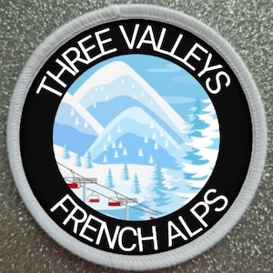 Op de afbeelding: Een witte en zwarte ronde patch met de tekst "Three Valleys French Alps" en een afbeelding van een besneeuwd berggebied met een skilift.