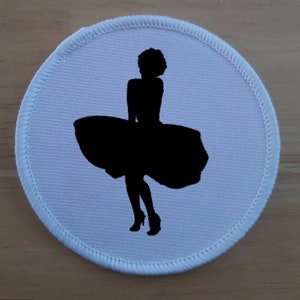Pode incluir: Patch redondo de tecido branco com uma silhueta preta de uma mulher vestindo um vestido com uma saia fluida.