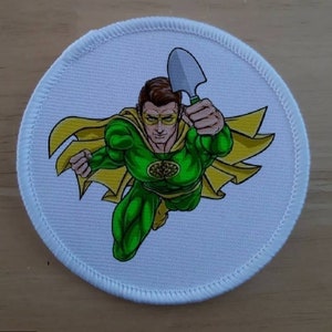 May include: A green superhero with a yellow cape and a small shovel flies through the air. The superhero has a tree logo on their chest.
