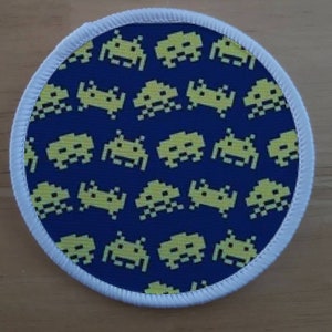May include: A round, white fabric patch with a blue background and a repeating pattern of yellow pixelated space invaders.