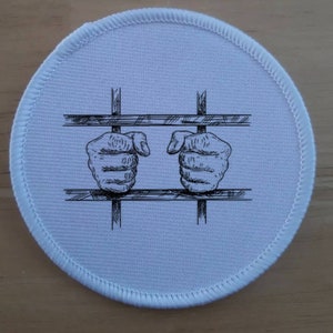 Puede incluir: Un parche redondo blanco con un borde cosido. El parche presenta una ilustración en blanco y negro de dos manos agarrando los barrotes de una celda. El diseño está centrado en el parche.