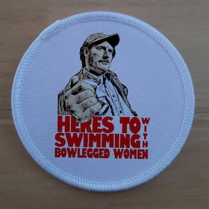 Pode incluir: Etiqueta redonda branca com borda vermelha e uma imagem de desenho animado de um homem com chapéu e camisa branca. O texto "HERE'S TO SWIMMING WITH BOWLEGGED WOMEN" está impresso em vermelho.