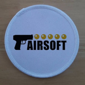以下が含まれることがあります： 黒い文字で「AIRSOFT」と書いてある白い丸いパッチ。黒い銃のシルエットと、文字の上に黄色い丸が6つある。