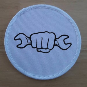 May include: White round fabric patch with a black outline of a fist holding a wrench.