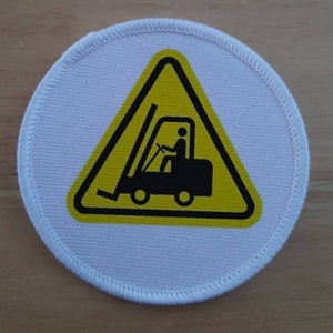 Peut inclure: Un écusson rond blanc avec un panneau de signalisation triangulaire jaune. Le panneau représente une silhouette noire d'un chariot élévateur avec une personne au volant.