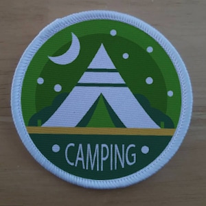 May include: A white camping tent with a green interior, surrounded by a green background with white stars. The text "CAMPING" is written in white on a yellow stripe below the tent.