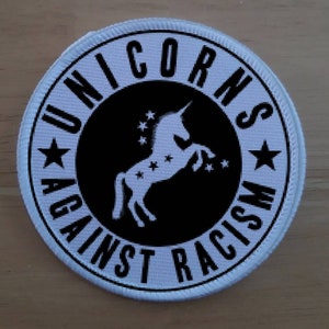 Può includere: Toppa rotonda bianca con testo nero e una silhouette di unicorno bianco. Il testo recita "Unicorns Against Racism" con una stella su entrambi i lati del testo.