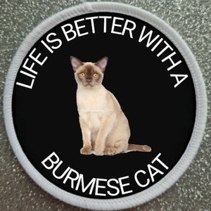 Op de afbeelding: Een ronde witte en zwarte patch met de tekst "Life is better with a Burmese cat" in witte letters. Een bruine Birmaanse kat zit in het midden van de patch.